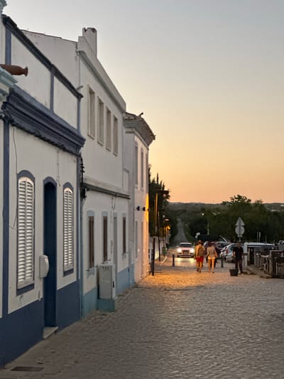 This image captures a view from a street in Vila Real de Santo António, Portugal, at dusk or dawn. The foreground shows a row of white buildings with blue trim, a common architectural style in the region. The buildings have multiple windows with shutters.
On the right side of the frame is a cobbled street, where several people are walking. Further along the street, cars are parked, and some are moving, their headlights visible. The background transitions into a hazy sky with a gradient of yellow and orange hues, suggesting either sunrise or sunset. Some trees are visible in the background.