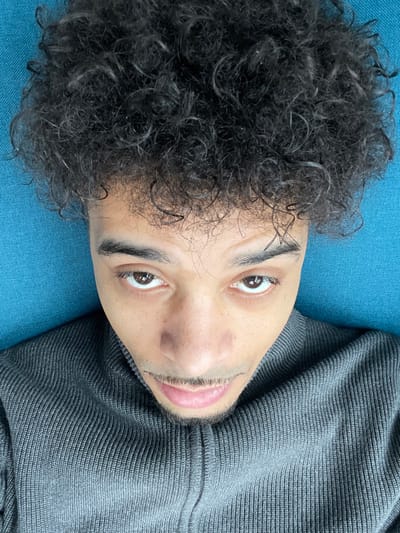 A single male individual is depicted in a close-up, high-angle shot, looking directly into the camera. He has dark, tightly curled hair and brown eyes. A thin mustache extends to a small goatee on his chin. He is wearing a grey, ribbed, full-zip turtleneck sweater. The background consists of a uniform, light blue, textured fabric surface, possibly upholstery. There are no other individuals or objects visible. The setting appears to be indoors. The image provides no visual information to confirm a location in Montigny-le-Bretonneux, France.