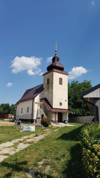 A vertical, medium-shot photograph captures a cream-colored church with a brown, multi-tiered steeple under a bright blue sky dotted with white clouds. The steeple is topped with a finial. The church has a dark brown roof, and a smaller attached structure also has a dark brown roof. There are windows visible on both the steeple and the attached structure. In front of the church, a stone cross stands in front of a rose bush. A pathway, made of flagstones and gravel, runs alongside a grassy area to the right of the church. In the foreground, a blue and white tarp covers some objects on the ground near a gravestone. The scene appears to be set in a rural or village environment, possibly a churchyard or cemetery. The sun is shining, suggesting it is daytime. The image is a still shot with no discernible activity taking place. There is no visible text in the image. The location is identified as Vyšné Nemecké, Slovakia.