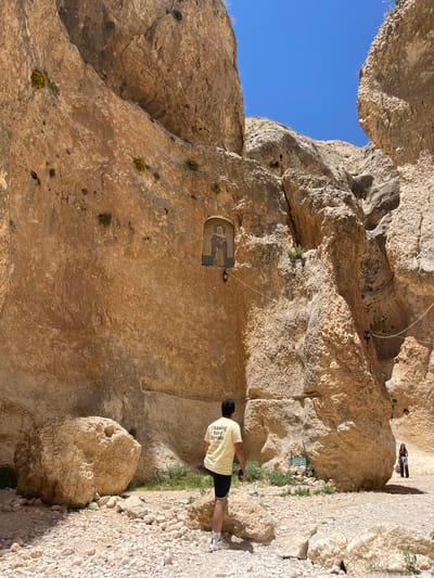 The image shows a partially rotated view of a rocky landscape with human presence. On the right, a man wearing a yellow t-shirt and dark shorts stands on a gravel path that runs along a rock face. An icon is embedded in the rock face. In the background, two figures are visible further up the path. Blue sky is visible through a gap in the rocks above. This is likely the Monastery of Saint Sergius and Bacchus in Ma'loula, Syria.