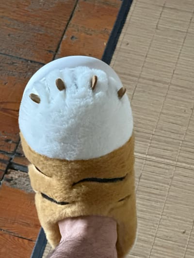 The scene is located in Annœullin, France. A human foot, visible from the lower ankle, is shown wearing a plush slipper. The slipper is predominantly tan/light brown with black stripes, featuring a white, fluffy toe area from which five brown, elongated, claw-like protrusions extend. The visible skin on the foot is light-toned and exhibits body hair. The floor beneath consists of reddish-brown wooden floorboards on the left and a light, straw-colored, ribbed mat on the right, separated by a dark, narrow linear element.