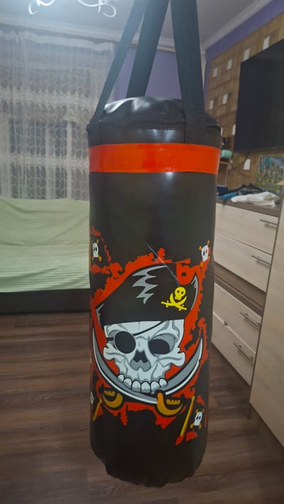 A black punching bag with a pirate skull and crossbones design is hanging in a room. The punching bag has red stripes at the top. In the background, there is a light-colored couch with a green blanket, sheer white curtains, a wooden floor, and a dresser with light-colored drawers. The overall scene suggests an indoor domestic setting.