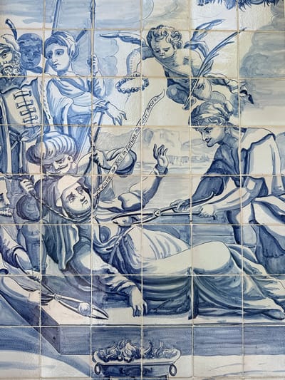 The image is a detailed close-up of a blue and white tiled mural depicting a scene with multiple figures and a central action. In the foreground, a person is lying down, appearing to be undergoing a procedure or examination, with medical instruments nearby. Another figure, possibly a medical practitioner, is leaning over the reclining individual, holding instruments. Above them, a cherubic figure, likely an angel, is depicted in flight, holding a scroll or ribbon with text. The mural shows a group of onlookers dressed in historical or regional attire, some holding spears or poles. The background is stylized, suggesting an architectural setting with buildings in the distance and a cloudy sky. The text on the scroll appears to be in Latin. The tiles are arranged in a grid, indicating they form part of a larger tiled artwork. The specific location of this mural in Mukhrani, Georgia, is not discernible from the visual information provided in the image itself.