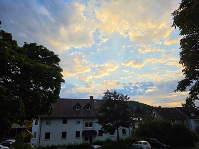 This image captures a sunset over a residential area in Höxter, Germany. The sky is filled with a mix of blue sky and dramatic clouds illuminated by the setting sun, casting golden and yellowish hues. In the foreground, dark green trees frame the scene, with their leaves silhouetted against the sky.
Below the sky, a multi-story building with a brown tiled roof dominates the midground. The building is painted white and has several windows, some of which appear to have occupants visible inside. Two dormer windows are prominent on the roof. A smaller tree with dark foliage stands in front of this building. Further back, other houses with similar tiled roofs are visible, nestled amongst trees and greenery. Several cars are parked in front of the buildings, including a white hatchback and a dark sedan. The overall impression is one of a peaceful evening in a suburban or semi-rural part of Höxter.