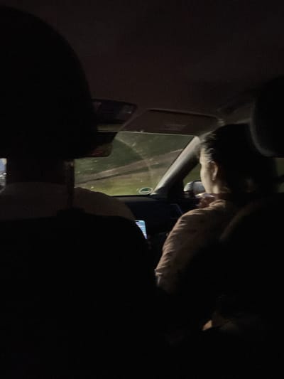 The image shows the interior of a car at night. Two individuals are visible, seated in the front passenger and driver seats, respectively. The driver, on the left, is largely obscured by darkness, with only the back of their head and shoulder visible. The passenger, on the right, is a woman with dark hair, seen from behind and to the side. She appears to be looking down, possibly at a mobile phone, which is illuminated by its screen. The interior of the car is dark, with ambient light coming from the exterior through the windshield and windows, suggesting a street or rural environment. The overall scene conveys a sense of movement, likely during a car journey. There is no specific visual information within the image to confirm the location of Ohlstadt, Germany.