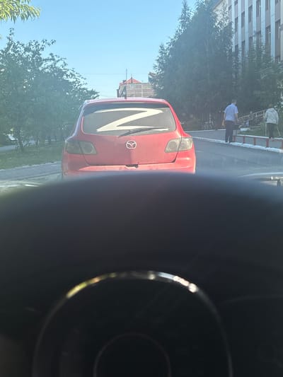The image captures a red Mazda car parked on a street in Nefteyugansk, Russia. The car's rear window features a prominent white "Z" symbol, a symbol that has been associated with the Russian military's invasion of Ukraine. The car is seen from behind, slightly to the left of the frame, with its taillights and Mazda logo visible. The street appears to be paved, with a sidewalk or pathway adjacent to it. In the background, a building with a distinct facade, possibly an office or residential complex, is visible. Several trees line the street, casting shadows and creating a somewhat shaded environment. Two individuals are walking on the sidewalk in the distance, one in a blue and white checkered shirt and the other in a gray hooded jacket. The sky above is clear and blue, indicating fair weather. The foreground of the image is dominated by the dark interior of a vehicle, likely the one from which the photograph was taken, with parts of the dashboard and steering wheel blurred.