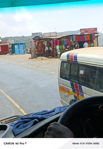 A street scene captured from the interior of a vehicle, showing the driver's hands on the steering wheel with a blue towel on the dashboard. Across the road, a small cluster of shops is visible. One sign reads "THE CLEAN TOILET", another "WELCOME TO VOLCANO CURIO", and a third "IMPALA CURIO SHOP." Goods such as animal hides and colorful fabrics are displayed for sale. A white van, labeled "KILGORISKLASSIC Sacco," is parked on the side of the road near the shops.