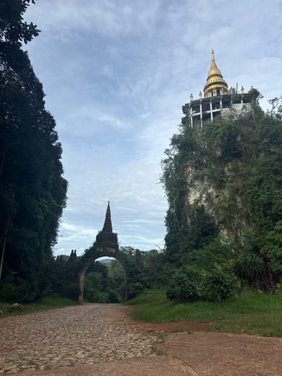 The image depicts the entrance to a temple complex situated in Ban Sadet, Thailand. A cobblestone path leads through an arched gateway adorned with two finials, one on either side of the arch. Beyond the gateway, a verdant jungle stretches out, with lush green trees forming a dense canopy. To the right of the path, a towering cliff face is covered in vegetation, with a golden-spired temple structure perched atop its summit. The sky above is partially cloudy, with a pale blue hue. The scene appears to be captured during daylight hours, likely in the morning or late afternoon, given the soft, diffused lighting. The overall atmosphere is serene and spiritual, evoking a sense of tranquility and natural beauty.