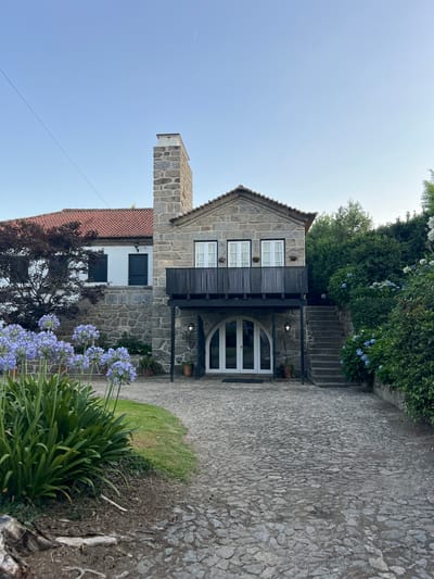 The image shows a stone house with a red-tiled roof in Felgueiras, Portugal. The house features a prominent stone chimney, a balcony with a black railing, and arched glass doors. In front of the house, there is a cobblestone driveway and a garden with blooming blue flowers. Lush green vegetation surrounds the property. The sky is clear and blue, indicating fair weather. There are no people or animals visible in the image. The scene appears tranquil and well-maintained, suggesting a residential or possibly a small guesthouse setting.