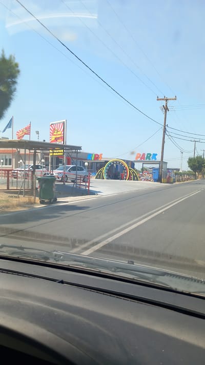 The image shows a street view of a "Kids Park" in Marmari, Greece, on a clear, sunny day. In the foreground, the dashboard and windshield wipers of a vehicle are visible, suggesting the photo was taken from inside a car. The road in front has white lane markings.

To the left of the road, a white building with a red roof is partially visible, with a covered area extending outwards. Two flags are displayed on poles in front of this building; one is blue, and the other is red with a design featuring a yellow figure. A large sign with red and white stripes and a yellow sunburst pattern, also featuring a yellow figure, stands near the building. Two white cars are parked under the covered area. A green trash bin is located near a metal fence.

Further to the right, a colorful archway stands, with the words "KIDS PARK" written in large, multicolored letters above it. The archway is decorated with what appears to be a speedometer design. To the right of the archway, there is a blue booth or structure. The ground in front of the archway is paved, and there are some colorful decorations or play structures visible.

In the background, utility poles and power lines stretch across the sky, which is a bright blue with some wispy clouds. Trees line the right side of the scene, and the horizon is mostly flat. The overall scene suggests a commercial establishment catering to children, located along a road in a coastal or semi-rural area.