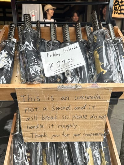 The image displays an outdoor retail vendor's stand in Chūō, Japan, featuring multiple black folding umbrellas designed with handles resembling samurai swords. These umbrellas are individually wrapped in clear plastic and arranged on two visible wooden display shelves. A white handwritten sign, centrally placed on the upper shelf, indicates "Folding Umbrella" and a price of "¥ 2700". Directly below this, a handwritten cardboard sign attached to the front of the upper shelf states: "This is an umbrella. not a Sword, and it will break. so please do not handle it roughly. Thank you for your cooperation." In the blurred background, three individuals are partially visible. The leftmost person wears a light cap and grey shirt, smiling and looking to the right. A second person with dark hair is visible in the center, and a third person with dark hair is on the right, both appearing to observe the surroundings. The scene is illuminated by natural daylight.