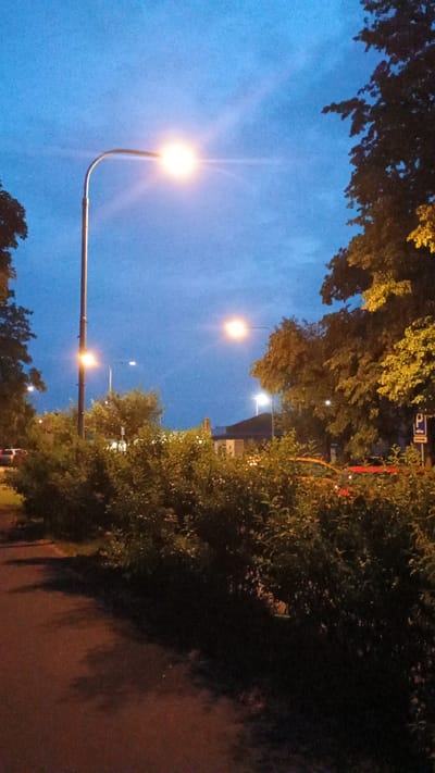 The image depicts an outdoor scene at dusk or dawn, likely in Sokolov, Czechia. The sky is a deep blue, transitioning to lighter shades as it meets the horizon. Streetlights are illuminated, casting a warm glow. On the right side of the image, lush green trees and foliage line a path or road, their leaves catching the ambient light. The shadows of the trees are cast onto the ground, suggesting a low light condition.
In the foreground on the left, a curved lamppost with a bright, glowing light is prominently featured against the sky. Further in the background, more streetlights are visible, some emitting a white light and others a warmer hue. Trees with yellow-tinged leaves are visible in the upper part of the image, indicating the presence of deciduous trees, possibly during autumn or late summer.
The scene appears to be a public space, such as a park or a street lined with trees. There are subtle hints of man-made structures in the background, partially obscured by the trees and foliage, but no clear identifying details are discernible. The overall mood is serene and peaceful, with the gentle illumination of the streetlights creating a tranquil atmosphere. No people are clearly visible in the image. There is no visible text in the media file.
