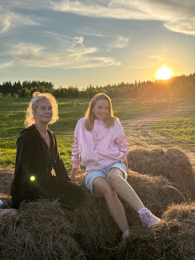This image depicts two women in a rural field during the golden hour of sunset. The bright sun, positioned low on the left side of the frame, casts a warm, golden light across the scene, creating a strong backlight that illuminates the subjects and causes a lens flare. The sky above is a clear blue adorned with wispy white clouds.

In the foreground, a woman with light, curly hair and a dark (possibly black) long-sleeved top is seated, looking directly at the camera with a relaxed, pleasant expression. Her hair is brightly backlit by the setting sun, giving it a luminous halo effect. Her light pink shoes are partially visible. Behind her and slightly to the right, another woman with long, straight blonde hair is reclining on a large, round hay bale. She is wearing a pink long-sleeved top and light blue denim shorts, with light pink sneakers visible on her feet. She is also looking towards the camera with a gentle smile.

The setting is an open field with green grass, leading towards a line of dark trees that form the horizon. The large hay bale occupies a significant portion of the right side of the frame, adding to the agricultural character of the scene. The overall atmosphere conveyed is peaceful and relaxed, capturing a moment of enjoyment in nature. There is no visible text in the image. This scene is located in a field near Стеклянный, Russia.