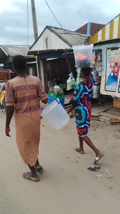 The image depicts an outdoor scene on a sandy path, likely within the city of Agbarho, Nigeria. Two individuals are walking away from the camera. The person on the left wears a long, light brown dress with horizontal darker brown stripes. They are carrying a large, translucent white plastic bucket with a blue handle in their left hand. The person on the right wears a colorful, knee-length dress with a pattern of blue, red, and white swirls. This individual is balancing a clear plastic container, filled with dark items and a bright orange object, on their head. In the background, there are several small, corrugated metal structures with white and yellow striped fronts, appearing to be market stalls or shops. Various goods, including crates of bottled beverages, bags of merchandise, and some food items, are visible outside these structures. The sky is overcast. The overall environment suggests a market or commercial area in a Nigerian town.