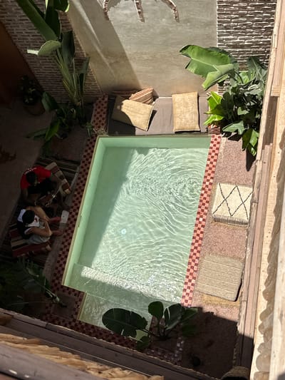 Overhead shot of a rectangular pool in Mechouar Kasbah, Morocco. The pool's edge is tiled with alternating red and tan squares. The pool is surrounded by various surfaces including tile, carpet, and wood. Two people are sitting poolside on a striped rug, one wearing a red shirt with white lettering and the other with a white towel around their head. Plants are located near the pool, including banana trees. Cushions are stacked by one side of the pool.