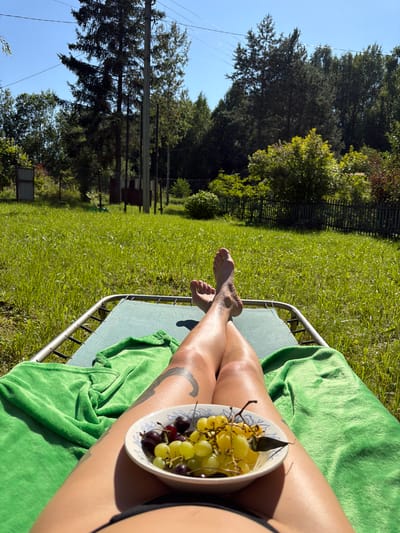 A first-person perspective shot shows a person lying on a green lounge chair outdoors, with a bright green blanket draped over their legs. A white bowl filled with green and red grapes sits on the blanket. The person's legs and feet are visible, with tattoos on their left calf and foot. In the background, a lush green lawn extends towards a wooden fence and a line of trees under a clear blue sky. Power lines are visible across the top of the frame. The setting appears to be a rural or suburban outdoor space, possibly a dacha or backyard, consistent with a location in Энтузиаст Снт (петрино), Russia.