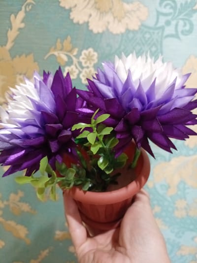 A hand holds a small terracotta pot containing two artificial flowers and green plastic leaves. The flowers have layered petals, transitioning from deep purple at the base to lighter shades of purple and then white at the tips. The background is a wall covered in wallpaper with a repeating pattern of stylized floral motifs in muted cream and blue-green tones. The lighting appears to be indoor, with no indication of time of day or weather. The scene is static, with no discernible activity taking place beyond the presentation of the potted flowers. There is no visible text in the image. The location is identified as Bologoe, Russia.