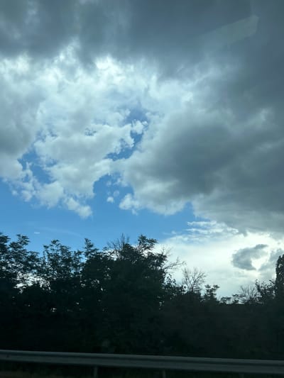 The image displays an outdoor scene in Dinkata, Bulgaria. The upper two-thirds of the frame are occupied by a sky featuring a mixture of bright white cumulus clouds, darker grey stratocumulus clouds, and visible patches of clear blue sky. The lighting indicates daytime. The lower third of the image consists of a dense, dark band of deciduous and evergreen trees and foliage, silhouetted against the sky. At the bottom of the frame, a horizontal grey metallic guardrail is visible. No individuals or specific actions are present within the scene. The perspective suggests the photograph was taken from a roadside or vehicle.