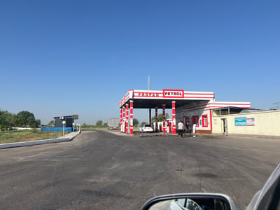 This is a frontal, eye-level, medium shot of a gas station in the city of Urustepa, Uzbekistan. The gas station has a white canopy with red trim, supported by red and white striped pillars. The canopy is labeled "PROPAN PETROL". There are several gas pumps under the canopy. A white car is parked at one of the pumps. Two individuals, one in a white shirt and black pants, the other in a dark shirt and black pants, are walking away from the canopy. Another individual in white is standing near the building. The gas station building is white with red trim around the windows and doors. A large poster is on the wall of the building, with the word "AYOQSH" visible. To the left of the gas station, there is a road leading away, with a large truck in the distance. The environment is dry and appears to be a rural or semi-rural area, with some trees and sparse vegetation visible in the background. The sky is clear and blue. The image is taken from the perspective of someone inside a vehicle, as evidenced by a car mirror in the foreground.