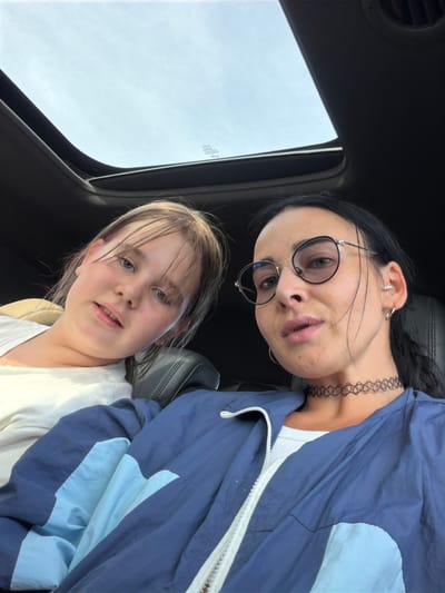 Two individuals are inside a vehicle, with the car's sunroof open above them. The person in the foreground, a woman with dark hair, wears round glasses, a black choker necklace, and a blue and light blue jacket over a white top. To her left is a younger individual, likely a girl, with light brown hair and blue eyes, wearing a white t-shirt. The background shows the interior of the car, including black car seats and a glimpse of the sky through the sunroof. The scene is set in a public place in the city of Sholokovo, Russia.