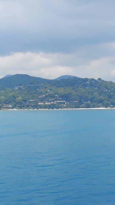 The image shows a coastal scene, likely in Baan Nai Wok, Thailand, based on the presence of lush, green vegetation on a hillside, beachside properties and the clear blue water. The scene includes a mountain covered in greenery on the left side and the calm sea on the right. There is visible evidence of sparse buildings within the trees, which indicates the area has properties. It is a sunny day, with few clouds.