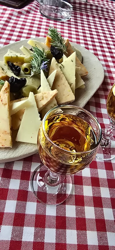 A top-down medium shot depicts a charcuterie board and two stemmed glasses filled with golden liquid, likely a type of wine or spirits, placed on a red and white checkered tablecloth. The charcuterie board contains an assortment of triangle-cut cheeses, olives, and herbs, seemingly rosemary, arranged artistically on a wooden tray. The glasses are positioned to the right of the board, partially overlapping its edge. The background consists of the red and white checkered tablecloth, which provides a vibrant contrast to the colors of the food and drink. There are no people visible in the image, and no specific location within Kopito, Montenegro can be determined based on the provided visual information. The overall scene suggests a casual dining or snack setting, possibly outdoors.