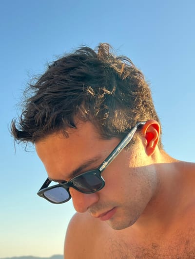 This image depicts a male individual, shown from approximately the upper chest to the top of the head, against a clear blue sky background, presumably in Agios Ioannis Diakoftis, Greece. The subject's head is tilted downwards, with the face largely obscured by dark-framed sunglasses. The sunglasses feature a patterned or mottled temple arm. Dark, dishevelled hair is visible, appearing somewhat windblown. The subject's skin exhibits a warm, tanned hue, consistent with exposure to direct sunlight, which illuminates the scene from the right, casting a bright sheen on the hair and skin. A portion of the right ear is visible, holding the sunglass temple arm in place. The upper torso, including a shoulder and part of the chest, is visible, suggesting a shirtless state, with visible body hair on the chest. No other individuals or significant objects are discernible. The environment consists solely of a bright, cloudless sky, implying an outdoor setting during daylight hours.