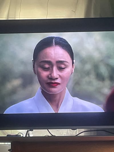 This media file shows a close-up, head-and-shoulders shot of a woman. She is wearing a white garment, possibly a traditional dress, with a high neckline. Her dark hair is slicked back and parted in the center. She has red lipstick on and her eyes are closed, giving her a serene or contemplative expression. The background is softly focused, appearing to be an outdoor scene with muted green and gray tones, possibly suggesting foliage or a natural landscape. The lighting suggests it could be daytime, but the overall mood is subdued. The image is presented in a vertical orientation, as if it is a still from a film or television program. There is no visible text in the image. The setting is implied to be in Linëvka, Russia, though no specific visual cues within the image directly confirm this location.