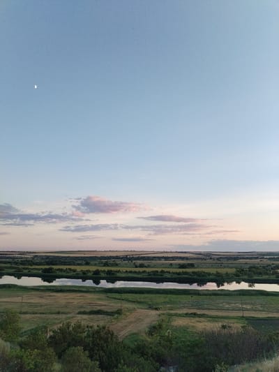 This is a landscape photograph taken in Bilousivka, Ukraine, likely during the late afternoon or early evening. The sky is a pale blue with a few wispy clouds tinged with pink and light purple, suggesting the approach of sunset. A small crescent moon is visible in the upper left quadrant of the image.
The scene depicts a rural panorama featuring a wide river or lake that reflects the sky and the trees lining its banks. The water appears calm and still. Beyond the water, rolling hills are covered in fields of crops, some appearing golden and harvested, while others are still green. Patches of trees and shrubs dot the landscape, creating a sense of depth.
In the foreground, there are grassy slopes and areas of tilled fields, with some scattered vegetation and what appear to be utility poles. The overall impression is one of natural beauty and tranquility in a rural setting. There are no people or obvious man-made structures, emphasizing the natural elements of the landscape. The lighting and colors evoke a peaceful and serene atmosphere.