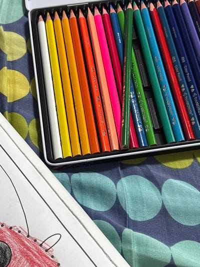 The image captures a close-up, elevated view of art materials situated on a patterned textile surface. The primary subjects are an open metal tin containing a collection of colored pencils and a portion of a white coloring page.

The metal tin, white on the exterior with a black plastic or foam interior insert, holds approximately 25 colored pencils. These pencils are arranged in rows, displaying a spectrum of hues including white, various yellows, oranges, reds, pinks, greens, and blues, extending to dark blue and purple. Several pencils are branded "TIANBOWEN" and labeled "4M Color Pencil." One green pencil lies diagonally across other pencils within the tin, disrupting the otherwise orderly arrangement.

To the lower left of the tin, a section of a white paper sheet, consistent with a coloring page, is visible. This page features black outline drawings. A specific area of the drawing, depicting part of an object (possibly a vehicle), has been colored in red, exhibiting a textured application of the coloring medium, along with small black circular elements.

The underlying surface is a fabric, possibly bedding or a blanket, characterized by a pattern of large, rounded, irregular shapes in teal and lime green/yellow set against a dark blue background.

No individuals are present in the frame. The arrangement of the items indicates an active or recently completed art activity. The precise location in Kafr Bara, Israel, cannot be ascertained from the visual evidence within the image.