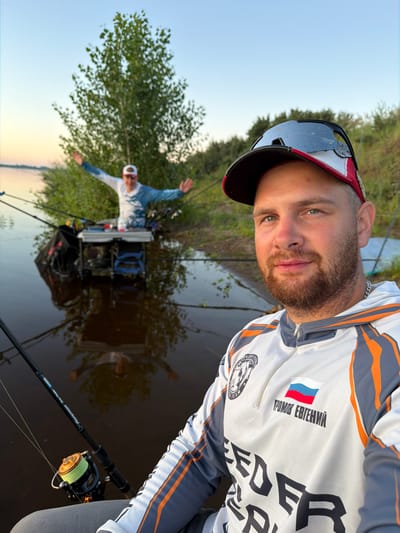 In the city of Tarasovo, Russia, two individuals are depicted near a body of water, likely engaged in fishing. The foreground is dominated by a man in a white, grey, and orange fishing jersey, wearing a baseball cap with sunglasses perched on top. He has a beard and is looking towards the camera. The jersey features Russian flag insignia and text. In the background, another person stands with arms outstretched, wearing a similar fishing outfit and a cap, facing away from the camera and towards the water. They are positioned behind a fishing station equipped with rods and tackle. The scene is set by a calm body of water reflecting the sky and surrounding trees and vegetation. The lighting suggests it is either early morning or late evening.