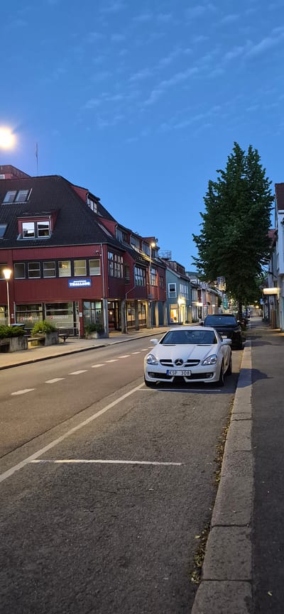 The image captures a street scene in Lillehammer, Norway, during twilight. The sky is a deep, clear blue with a few scattered, wispy clouds. A bright street light illuminates the left side of the frame, casting a warm glow on a red-colored building. This building has a dark, sloped roof with multiple windows and dormers. A prominent blue neon sign on the building reads "TRAALLAUG".

In the foreground, a white convertible Mercedes-Benz SLK with license plate "KSP 308" is parked on the right side of the street. Behind it, a dark SUV is also parked. The street is paved with asphalt and marked with white lines indicating parking spaces and a lane divider. A gray concrete sidewalk runs along the right side of the street, with a curb separating it from the road. A tall, leafy green tree stands on the sidewalk, its branches reaching towards the sky.

Further down the street, other buildings with colorful facades (green and blue) are visible, some with illuminated windows suggesting businesses or residences within. The overall impression is a quiet, urban street scene as day transitions into night. There are no people visible in the image.