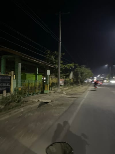 The image depicts a street scene at night in Tanjungkarang, Indonesia. The perspective is from a moving vehicle, likely a motorcycle, as evidenced by the presence of a side mirror in the lower right corner. The road is visible on the right side, appearing wet and reflecting the lights from vehicles and streetlights. On the left side of the image, there is a utility pole with numerous wires extending across the frame. Beyond the pole, a building with green fencing and what appears to be a dark-colored roof is visible. The building has multiple windows, some of which are illuminated. Further down the street, other vehicles and buildings are blurred due to motion and the low-light conditions. The overall impression is of a typical urban night scene, with the activity of traffic and the presence of infrastructure like utility poles and buildings. The time of day is clearly night, and the wet road suggests recent or ongoing rain. There is no discernible emotion conveyed, and any visible text is illegible due to blur and distance.