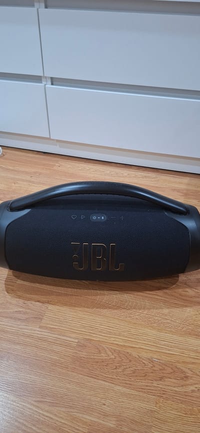 A black JBL portable speaker with a gold JBL logo is placed on a light brown wooden floor. The speaker has a black mesh front and a black handle. Above the JBL logo are several control buttons, including a power button and Bluetooth symbol. In the background, a white cabinet with horizontal drawers is visible. The scene appears to be indoors.