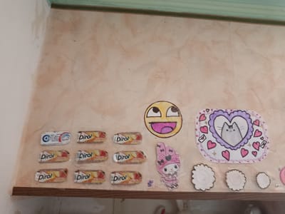 The image shows a collection of small, colorful drawings and candy wrappers arranged on a textured, peach-colored wall. The items are placed on a wooden shelf that runs along the bottom of the frame.
On the left side, there are seven candy wrappers, one white "Orbit" wrapper and six "Dirol" wrappers. Above the "Dirol" wrappers, there is a drawing of a smiley face with wide eyes and a large open mouth. To the right of the smiley face, there is a drawing of a grey cat within a purple heart, surrounded by pink hearts and envelopes. Below this, there is a drawing of a pink character with a bow and a dress, resembling a cartoon bunny. To the right of the cat drawing are three circular white cutouts, and further to the right, partially visible, is a pink circular drawing. A small blue drawing of a character is placed below the pink bunny.
The wall appears to be interior, with a green border along the top edge. The overall scene suggests a decorative arrangement or personal display. The location is identified as Dachne, Ukraine.