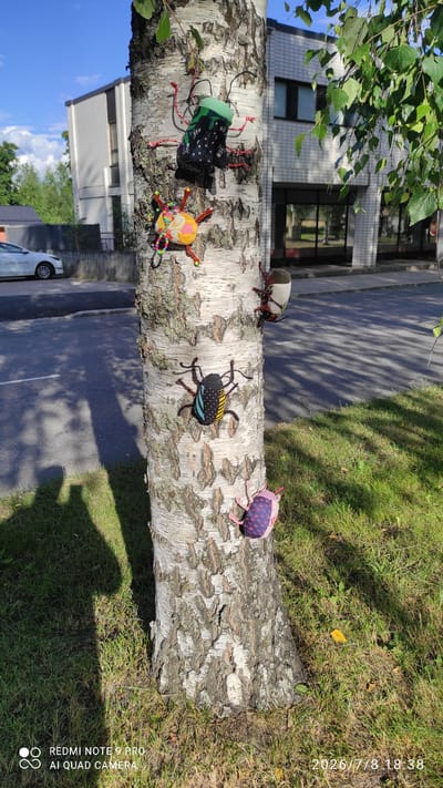 The image shows a fallen birch tree trunk, laid horizontally across a grassy area next to a paved path and a building. Attached to the trunk are five decorative, handmade insects.

The insects are varied in color and design. One has a green top and black fabric with a white pattern for its body, and red pipe cleaner legs. Another has a yellow and orange body with colorful beaded decorations and red pipe cleaner legs. A third is black with yellow and blue polka dots and black pipe cleaner legs. The fourth is gray and white with red pipe cleaner legs. The fifth is covered in a pink and purple patterned fabric with pink pipe cleaner legs.

The scene appears to be outdoors, likely in a park or residential area, given the presence of the building and paved path. Green leaves from a nearby tree hang into the upper left of the frame. The lighting suggests it is daytime, with shadows cast on the grass and path. The sky is blue and partly cloudy.

Visible text in the image includes a watermark in the lower right corner that reads "REDMI NOTE 9 PRO AI QUAD CAMERA" and a timestamp "2026/7/8 18:38" on the right side. The location context provided is Iisalmi, Finland.