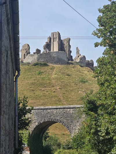 The image depicts the ruins of Corfe Castle situated on a grassy hill under a clear blue sky. In the foreground, a stone bridge with an arched opening spans across the lower part of the frame. To the left, the weathered stone wall of a building runs vertically. Lush green trees frame the right side of the image, with some foliage also visible beneath the bridge. A pathway is visible winding up the grassy slope towards the castle ruins. Electrical wires are suspended across the sky. The overall scene is a picturesque view of historical ruins and natural landscape.