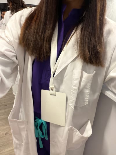 The image shows an overhead view of a person wearing a white lab coat over a vibrant purple uniform or scrubs. The person's long, dark brown hair flows down their back and shoulders, partially obscuring the lab coat. A white, rectangular ID badge holder with a strap is clipped to the left side of the lab coat. Peeking out from under the lab coat on the right side is a tied teal ribbon or string, possibly belonging to another part of the uniform or an accessory.
In the bottom left corner, a second person is partially visible, wearing a white t-shirt. Text is visible on this t-shirt, appearing to read "UPAF" followed by obscured letters. The background suggests an indoor setting, with a light-colored wall or surface and a wooden floor visible in the lower right portion of the image. The overall scene depicts someone dressed in professional or educational attire, possibly in a medical or scientific environment.