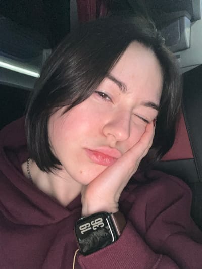 A woman with short, dark hair rests her head on her hand, partially closing one eye. She wears a maroon hoodie, and an Apple Watch is visible on her wrist, displaying the time 19:36. The lighting suggests an indoor environment. The background is dark, and the seating appears to be in transit. The overall image conveys a sense of weariness or relaxation.
