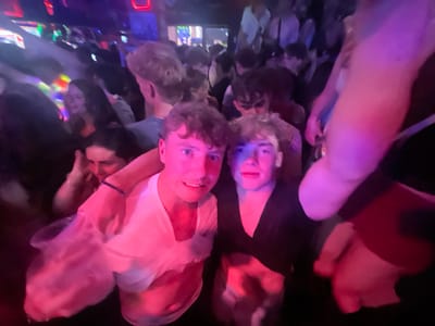 The image depicts a crowded indoor environment, consistent with a nightclub or bar located in Albufeira, Portugal. The scene is characterized by low light conditions and saturated artificial illumination, primarily in red, blue, and purple hues.

In the foreground, two young adult males are centrally positioned, looking directly at the viewer with slight smiles. The male on the left, with light, curly hair and wearing a white t-shirt, has his left arm placed around the shoulder of the male to his right. His right hand appears to hold a translucent plastic cup. The male on the right, also with light, curly hair, is wearing a dark top.

The background is densely populated with numerous other individuals, largely out of focus due to the depth of field and motion blur, indicating a high concentration of people. Among them, a female with dark hair is visible behind the two main subjects, her hands near her face. The overall environment suggests an active social gathering with typical nightclub characteristics, including dynamic lighting and a dense crowd. Distant light sources and indistinct architectural elements are present in the upper background.