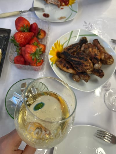 The image presents a table setting with various food items and a drink. A plate of grilled chicken pieces is visible in the upper center, next to a white plate and a fork. A container of strawberries sits on the lower left. A partially filled glass with a yellow liquid, possibly sparkling wine or champagne, is in the lower right corner. Foil-wrapped items are placed under the glass. Other food items are partially visible on a plate in the left part of the image. The background is a white tablecloth with a subtle floral pattern. The photo provides no evidence of location in Bashur, Russia or information about people.