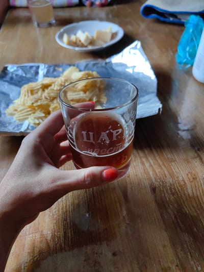 A hand with red painted fingernails holds a glass of amber liquid with white lettering. The glass is partially filled, and condensation is visible on the exterior. Behind the glass, a pile of ridged potato chips is spread on a piece of crinkled aluminum foil. A white plate with cubed cheese is visible further in the background, along with a glass of a lighter colored beverage. The scene is set on a wooden table with a visible grain pattern. The overall impression is that of a casual snack or meal. The location is specified as Фаніпальскі сельскі Савет, Belarus.