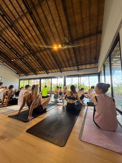 The scene depicts a group of approximately 20-25 individuals participating in a fitness or yoga class within a studio in Munggu, Indonesia. The room features a high, vaulted ceiling constructed from light-colored bamboo panels supported by dark metal beams, equipped with two dark-bladed ceiling fans, one visibly rotating, and multiple recessed light fixtures. The floor is light-colored wood. Large, floor-to-ceiling glass windows or sliding doors line the right wall, offering a view of dense green foliage and a distant building or structure.
The participants, primarily women, are seated on individual yoga mats arranged in rows, mostly facing away from the camera towards the front of the room. They are dressed in athletic attire, including sports bras, tank tops, and leggings or shorts. Visible objects include black yoga mats, some branded "HOISSIULU," and a pink mat branded "FITTASTIC." Several individuals have water bottles, small white towels, or yoga blocks positioned near their mats. The overall environment suggests a contemporary studio setting with natural and artificial lighting.