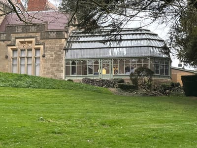 The image displays an exterior view of a stone building connected to a glass conservatory, situated on an upward-sloping green lawn in Malvern Hills, United Kingdom. The main structure, occupying the left, is constructed of light-colored cut stone blocks, featuring multi-paned windows with elaborate stone mullions and decorative carvings above. A section of a brown-tiled roof is visible.

To the right, an attached, multi-faceted glass conservatory features a green metal framework and a segmented glass roof. Numerous transparent window panes form its walls, through which internal illumination is visible. A set of green double doors with a pointed arch design is centered on one of the conservatory's facets. The conservatory rests on a low, rough stone retaining wall, fronted by various small shrubs and plants.

The foreground is dominated by a neatly mown, bright green grass lawn that slopes gently upwards towards the structures. Bare tree branches partially obscure the upper portions of the buildings and an overcast, light grey sky. In the far right background, a section of a tan-colored building with a dark roof and chimney, along with a trimmed hedge, is visible. No individuals or active interactions are present in the scene.