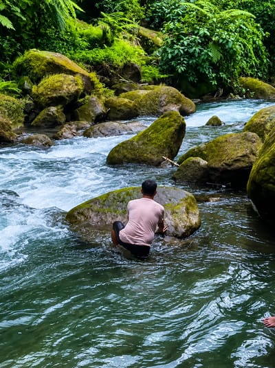 A person wearing a light pink t-shirt and black shorts is crouched on a moss-covered rock in a rushing river. The river flows over rocks, some of which are covered in moss. Lush green vegetation and trees surround the river, suggesting a natural, possibly jungle, environment. The water is clear with visible ripples and currents. A small portion of another person's feet is visible in the lower right corner of the frame, partially submerged in the water. The location is described as Gunung Ambat, Indonesia.