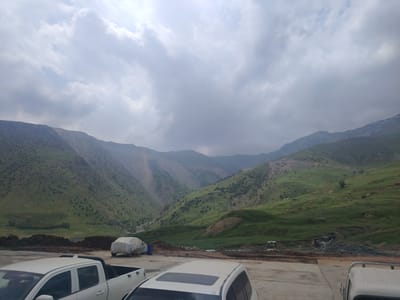 The image depicts a mountainous landscape with a cloudy sky. In the foreground, there are several vehicles parked on a paved surface. A white pickup truck is prominently visible on the left, and the rear of a white SUV is in the center. A vehicle covered with a tarp is parked behind the pickup truck. The background features rolling green hills and mountains, with some evidence of construction or excavation work, including piles of dirt and what appears to be construction machinery. The location is identified as the city of Farab, Tajikistan. There are no people clearly visible in the image.