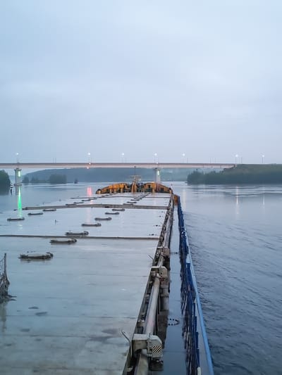 The image shows a view from the deck of a barge, moving forward on a wide river. The sky is overcast and the water is calm, with some ripples from the barge's movement. A long, modern bridge with streetlights spans the river in the distance, with treed banks visible on either side. The barge itself has a flat, grey deck with numerous rectangular hatches. The barge is blue on its starboard side. The overall scene suggests a quiet, early morning or late evening journey on a river, possibly near a populated area given the bridge. The location is identified as Ivanov Bor, Russia.