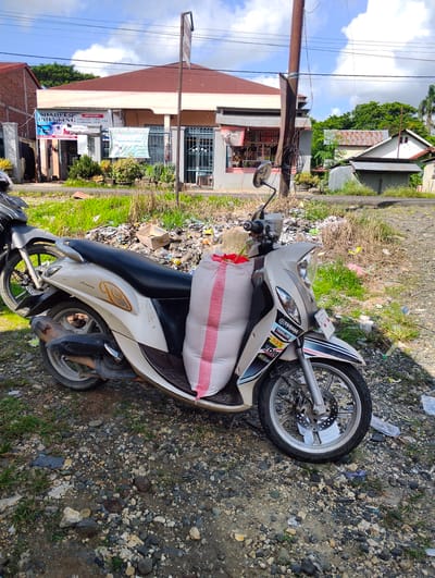 The image depicts a light-colored Yamaha scooter parked on a gravel and dirt surface in front of a building. The scooter has a large, white sack with red stripes tied to its rear seat. Stickers are visible on the front fairing of the scooter, including "YAMAHA" and "YAMALUBE". The building behind the scooter appears to be a commercial establishment with signs indicating printing services and a bimbel (tutoring center). In the background, there are other buildings, trees, and a power pole with wires. The ground is covered with scattered debris, including plastic bags, cardboard, and other refuse. The scene suggests an outdoor setting, possibly a street or a roadside area, in Pompanua, Indonesia. The sky is partly cloudy. There are no people visible in the image.