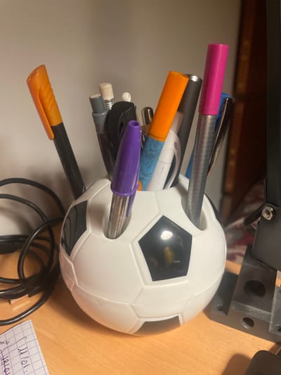 A soccer ball-shaped pen holder sits on a desk, filled with various pens and pencils. The holder is white with black pentagons. Pens of different colors including orange, pink, purple, and silver extend from the ball in different directions. The desk surface is light brown. A black cable and a small notepad are also visible on the desk. Parts of a black, mechanical object are visible in the top portion of the frame.