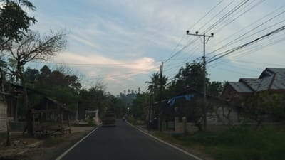 The image depicts a rural road in Luengbaro, Indonesia, during what appears to be dusk or early morning. The sky is a soft blue with wispy clouds. A paved road runs through the center of the frame, with a white line marking the edge. On the left side of the road, there is a small, open-air wooden shelter with a thatched roof, and a bare tree with intricate branches. Further down the road, on the left, are buildings with wooden and brick construction, and dense foliage. On the right side of the road, there are houses with corrugated metal roofs and some areas covered with blue tarps. A utility pole with multiple power lines stands tall on the right, casting shadows. In the distance, a truck is visible on the road, and a motorcycle can be seen further ahead. The landscape is characterized by lush green trees, some of which are palm trees, and a gentle incline of hills in the background. The overall impression is one of a quiet, developing rural community.